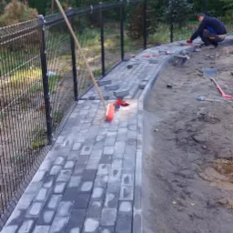 Układanie szarej kostki brukowej na posesji w Gdańsku, widoczny pracownik, narzędzia i częściowo ułożony chodnik wzdłuż ogrodzenia.