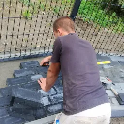 Układanie szarej kostki brukowej w ogrodzie, widoczny pracownik z tyłu, poziomica, gumowy młotek i ubijarka wibracyjna.