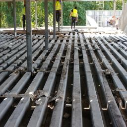 LAPIS HOME SP. Z O. O. - Demontaż starej podłogi z metalowych belek w budynku, widoczni pracownicy w odblaskowych kamizelkach, ślady gruzu i resztek materiałów budowlanych.