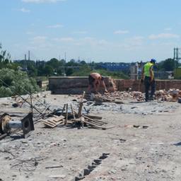 LAPIS HOME SP. Z O. O. - Demontaż ceglanej ściany na dachu budynku w słoneczny dzień, widoczni dwaj pracownicy w trakcie pracy, jeden bez koszulki, drugi w kamizelce odblaskowej, w tle zabudowa miejska i błękitne niebo.