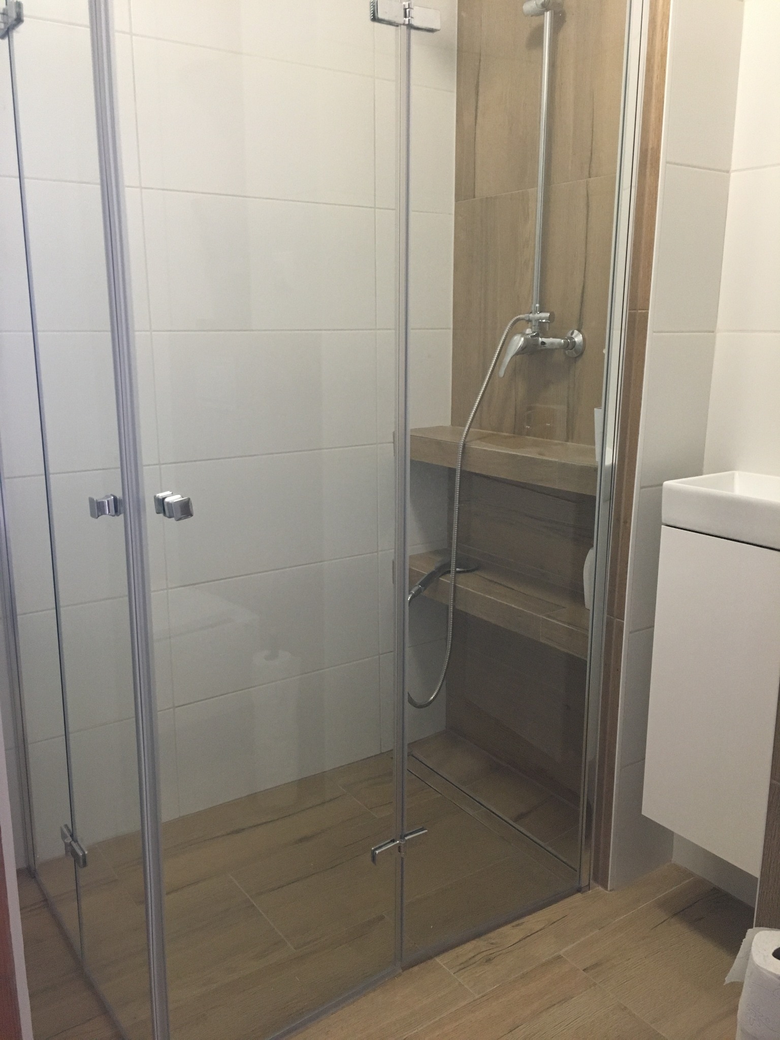 Nowoczesna kabina prysznicowa z szklanymi drzwiami i drewnopodobnymi płytkami w wyremontowanej łazience w Gdańsku, z widocznymi półkami wewnątrz kabiny i minimalistyczną umywalką.