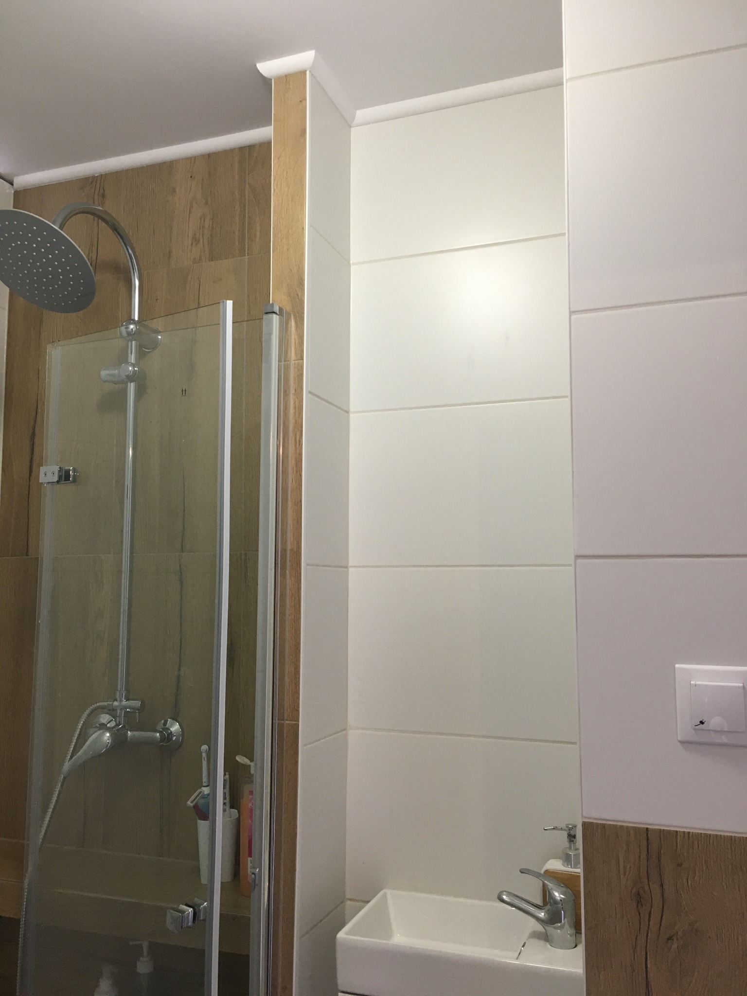 Nowoczesna kabina prysznicowa z deszczownicą i szklanymi drzwiami, obok umywalka z baterią i włącznik światła na białej ścianie z drewnopodobną okładziną.