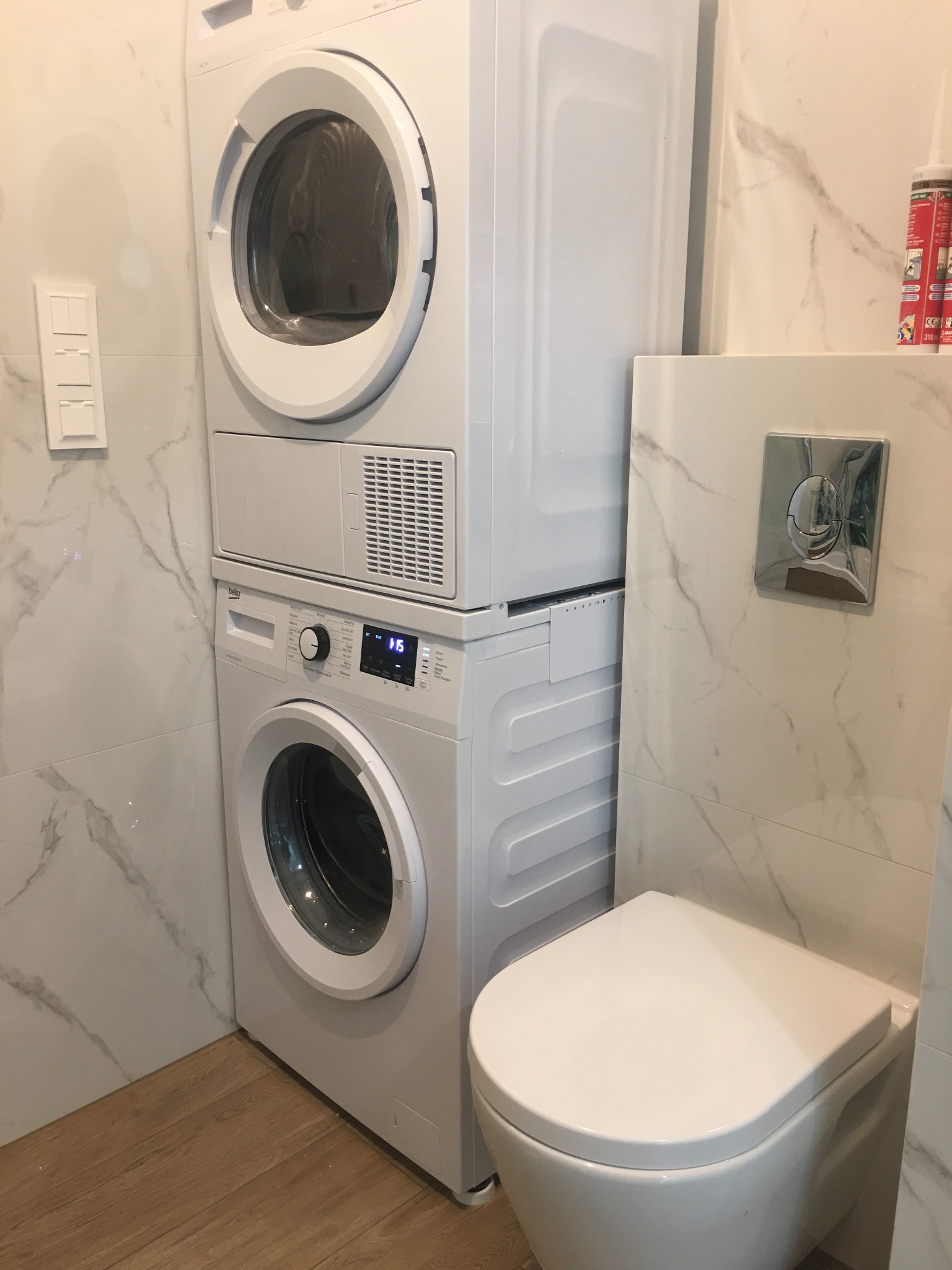Nowoczesna łazienka z marmurowymi płytkami, wiszącą toaletą i pralko-suszarką Beko ustawioną jedna na drugiej, podłoga wyłożona panelami imitującymi drewno.