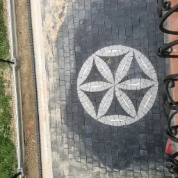 Ułożona kostka brukowa w geometryczny wzór kwiatu na czarnym tle, widok z góry, częściowo zabrudzona piaskiem, obok trawnik z żółtymi kwiatami i metalowe ogrodzenie.