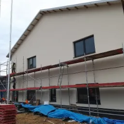 Ocieplanie elewacji budynku w Porażynie, widoczne rusztowania i pracownicy podczas nakładania warstwy izolacyjnej na ścianę, w tle ogrodzenie z drutu kolczastego.