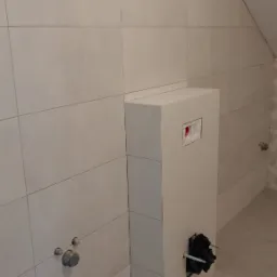 Ściana łazienki z beżowymi płytkami, zabudowa stelaża podtynkowego WC z widocznymi przyłączami hydraulicznymi i otworem rewizyjnym, poddasze.