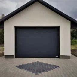 Nowoczesna brama garażowa segmentowa w kolorze antracytowym, z poziomymi przetłoczeniami, zamontowana w budynku z białym tynkiem i ciemnym dachem, wykończona kostką brukową z dekoracyjnym wzorem.