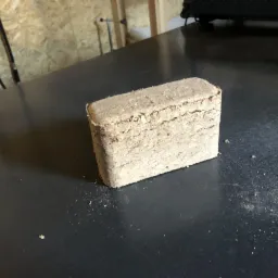 Prostopadłościenny brykiet drzewny z widocznymi warstwami sprasowanych trocin, leżący na ciemnym, matowym blacie. Drobne okruchy rozsypane wokół brykietu.