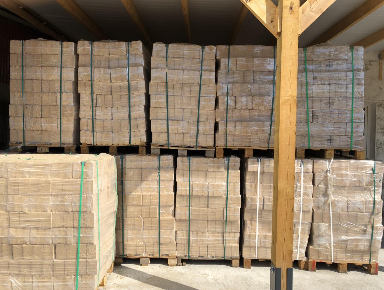 Magazynowane palety z brykietem drzewnym owinięte folią stretch, widoczne drewniane elementy konstrukcji dachu.