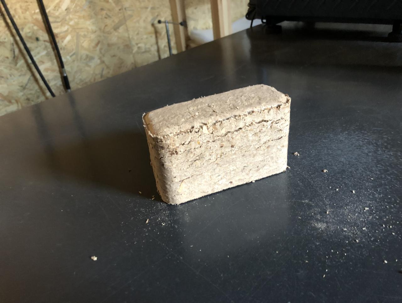 Prostopadłościenny brykiet drzewny z widocznymi warstwami sprasowanych trocin, leżący na ciemnym, matowym blacie. Drobne okruchy rozsypane wokół brykietu.