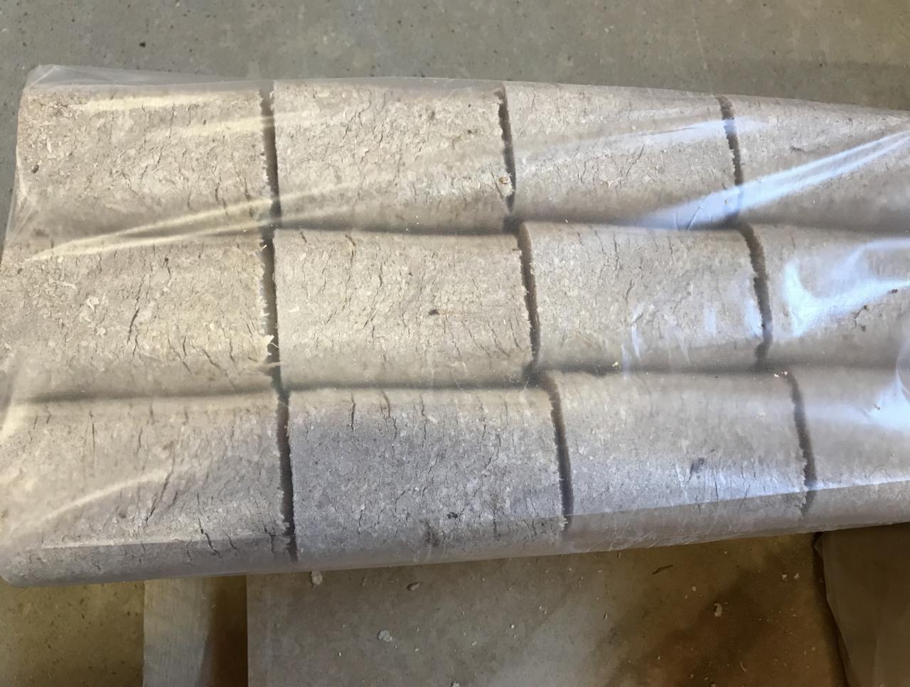 Rząd ułożonych cylindrycznych form paliwa opałowego z drewna, zapakowanych w przezroczystą folię, widoczne drobne pęknięcia na powierzchni.