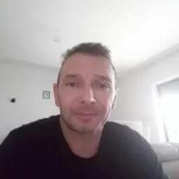 Portret mężczyzny w czarnej koszulce na tle jasnego pokoju z oknem i kanapą.