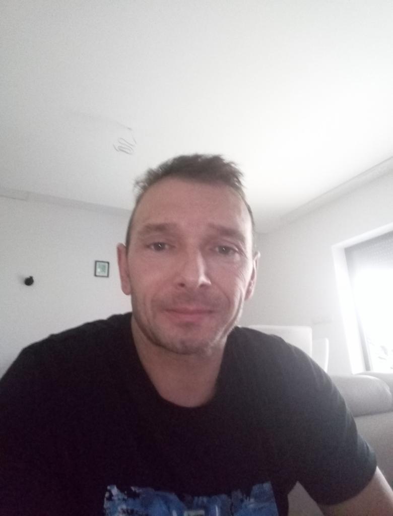 Portret mężczyzny w czarnej koszulce na tle jasnego pokoju z oknem i kanapą.