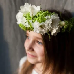 Portret dziewczynki z wiankiem z białych kwiatów i zielonych akcentów, spoglądającej w lewo, na tle szarej ściany.