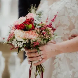 Panna młoda w koronkowej sukni trzyma bukiet ślubny z różowych, czerwonych i białych kwiatów, uchwycony z bliska, z widocznym pierścionkiem zaręczynowym.