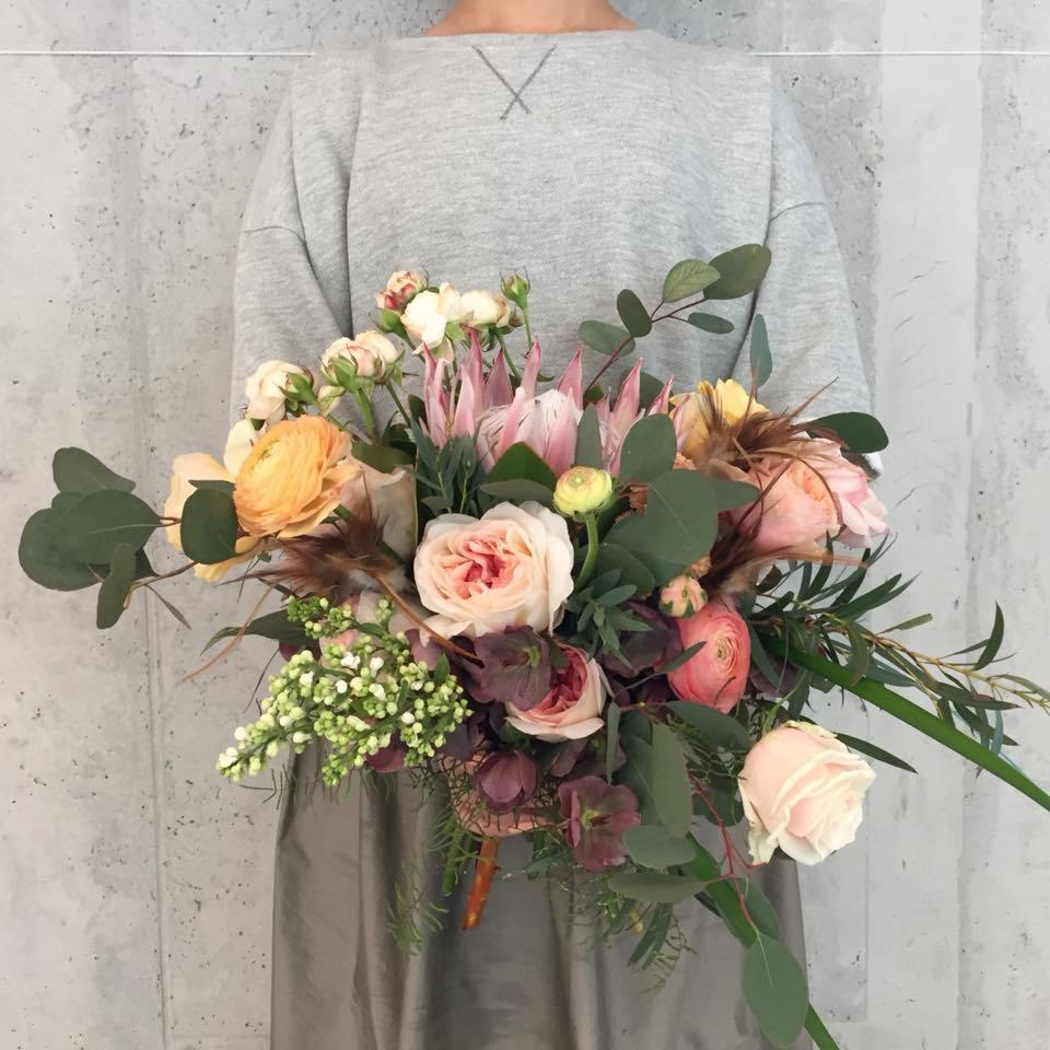 Osoba trzyma w rękach pastelowy bukiet kwiatów z róż, protei i eukaliptusa na tle szarej ściany.