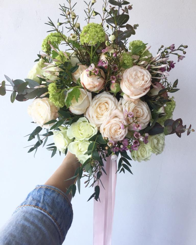 Delikatny bukiet ślubny z kremowych i zielonych róż, eukaliptusa i drobnych fioletowych kwiatów, trzymany w dłoni w dżinsowej kurtce, z długimi różowymi wstążkami.