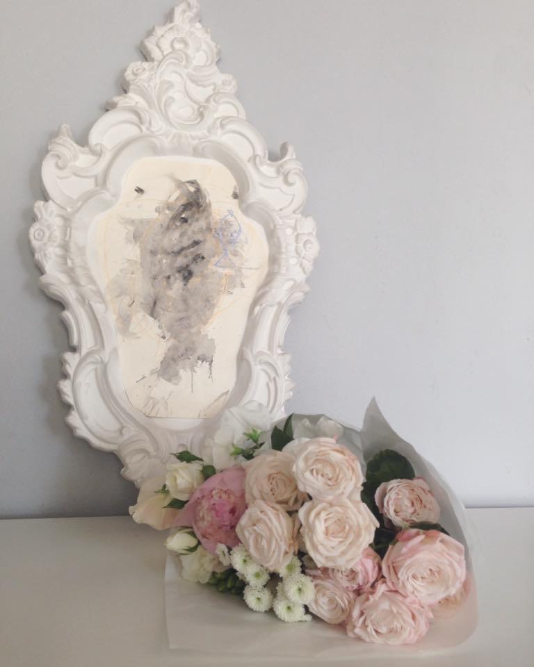 Biała, ozdobna rama z abstrakcyjnym malunkiem wewnątrz, obok bukiet różowych i białych kwiatów w białym papierze.