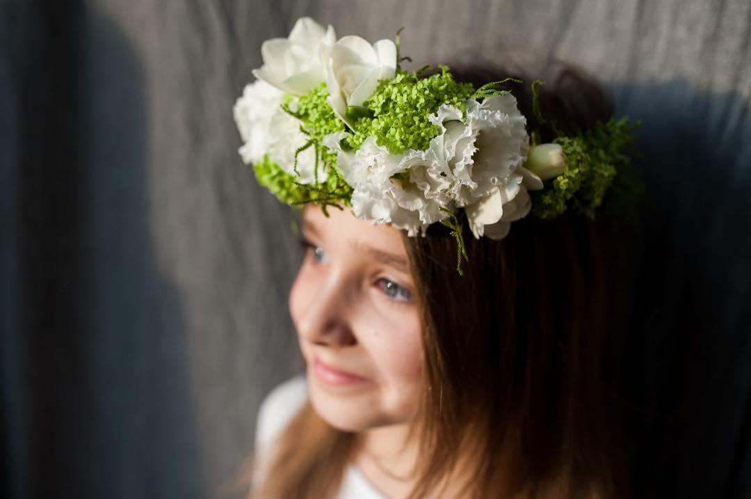 Portret dziewczynki z wiankiem z białych kwiatów i zielonych akcentów, spoglądającej w lewo, na tle szarej ściany.