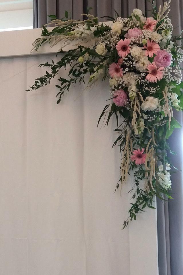 Dekoracja kwiatowa z różowych gerber, białych róż i zielonych liści, zdobiąca narożnik białej ściany z zasłonami w tle.