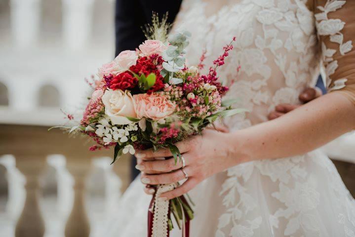 Panna młoda w koronkowej sukni trzyma bukiet ślubny z różowych, czerwonych i białych kwiatów, uchwycony z bliska, z widocznym pierścionkiem zaręczynowym.