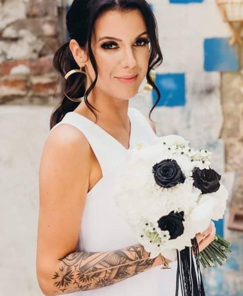 Uśmiechnięta panna młoda z tatuażem na ramieniu trzyma bukiet białych i czarnych róż na tle muru z niebieskimi akcentami.