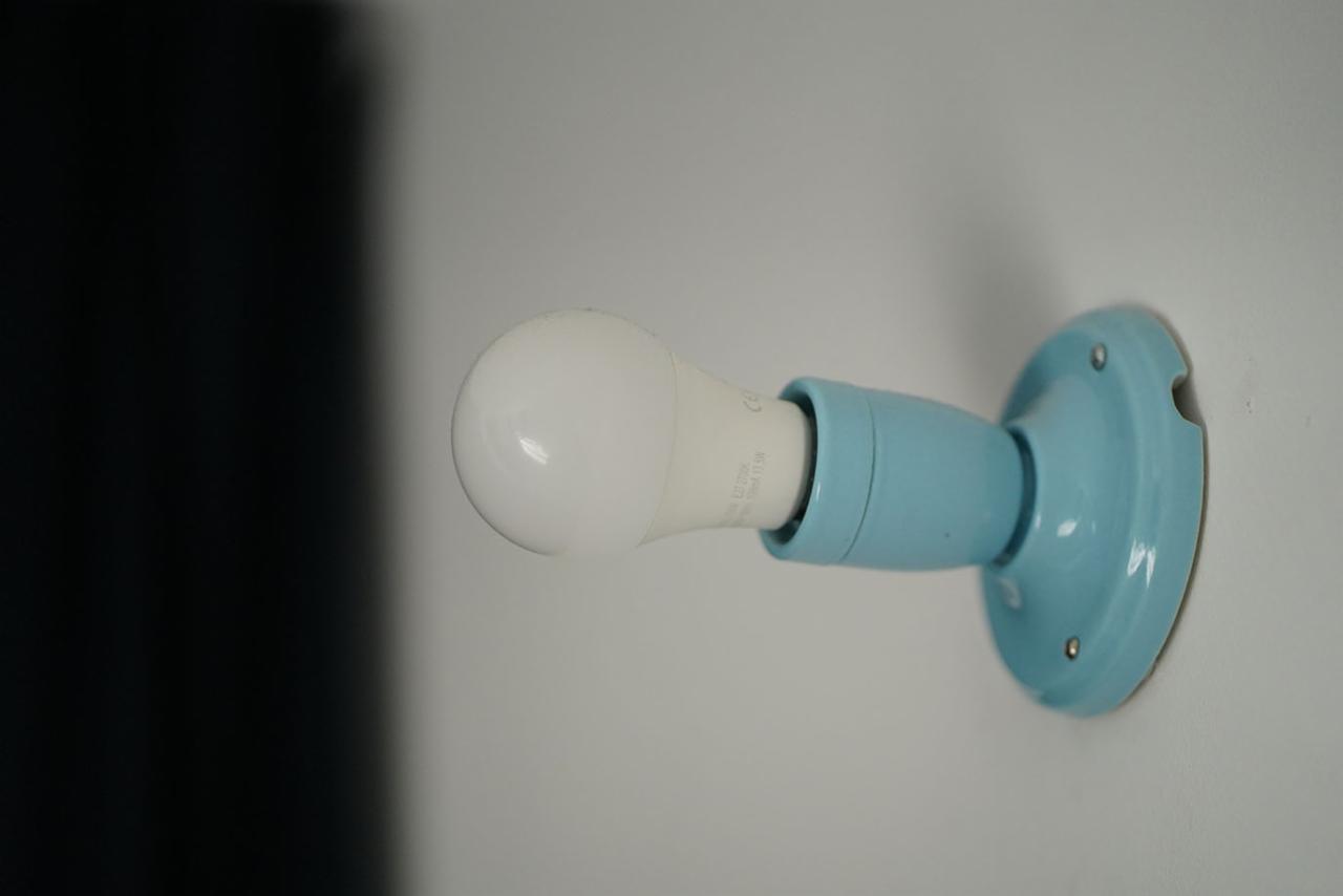 Biała żarówka LED w pastelowo błękitnej, ceramicznej oprawce zamontowanej na białej ścianie, z widocznymi śrubami mocującymi.