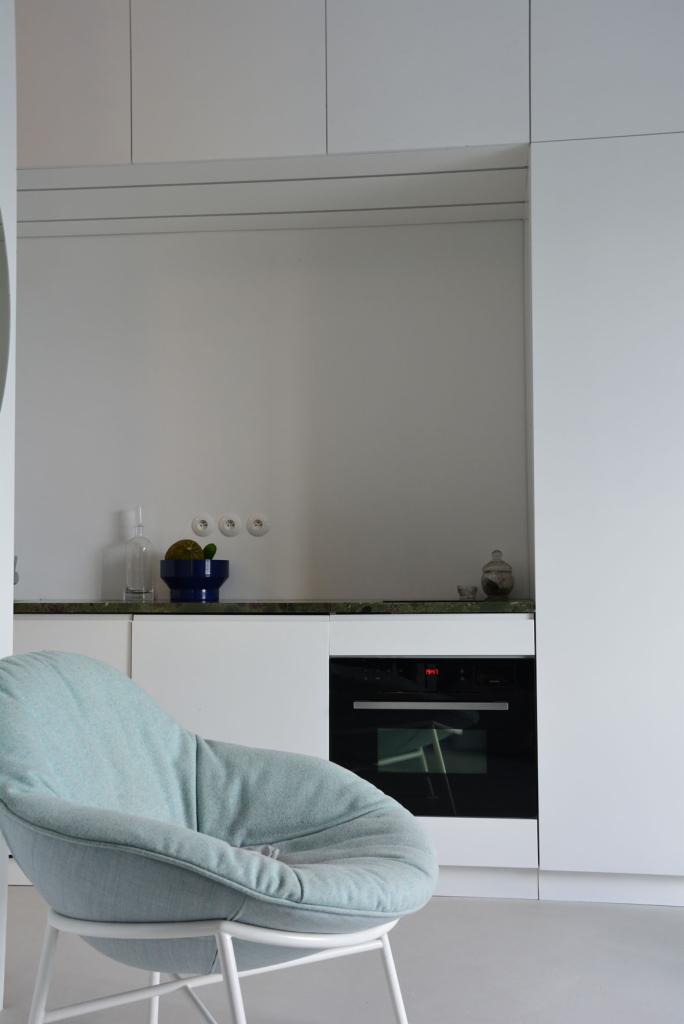 Minimalistyczna kuchnia w odcieniach bieli z zielonym blatem, z błękitnym fotelem na pierwszym planie, oświetlona naturalnym światłem.