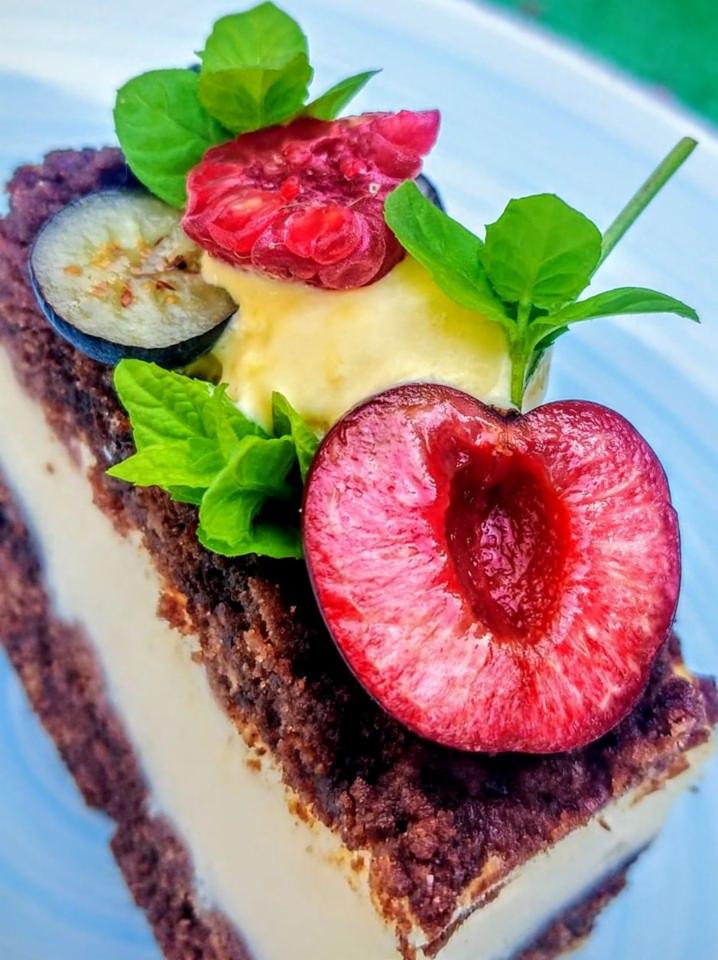 Kawałek ciasta z kremem, ozdobiony przekrojoną wiśnią, maliną, borówką i listkami mięty na jasnym talerzu, widoczne warstwy ciasta i kremu.