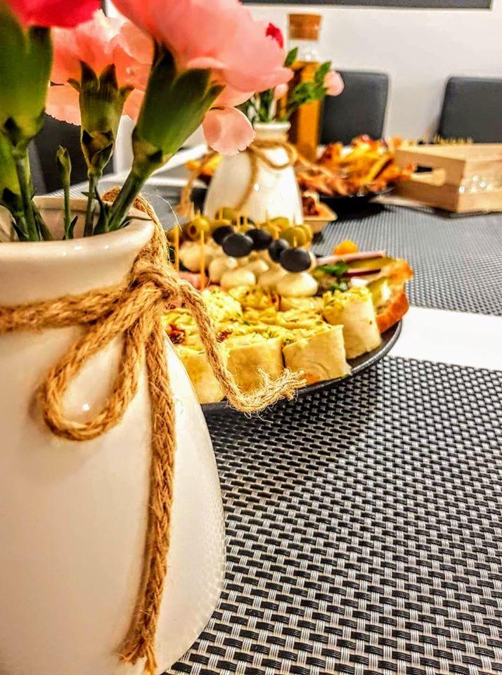 Udekorowany stół z cateringiem: rolki z farszem, koreczki z oliwkami i mozzarella, w tle kwiaty w wazonie z jutowym sznurkiem.