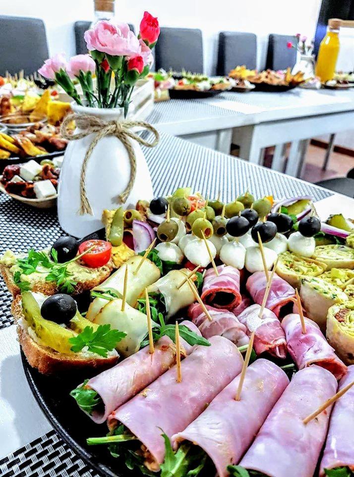 Elegancko przygotowany stół z różnorodnymi przekąskami: kanapeczki, roladki z szynki, koreczki z oliwkami i mozzarellą, oraz wazon z różowymi goździkami jako dekoracja.