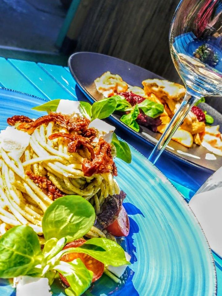 Dwa talerze z jedzeniem na niebieskim stole: pierwszy z makaronem spaghetti z pesto, suszonymi pomidorami i serem, drugi z pieczonymi ziemniakami, burakami i zieleniną. W tle kieliszek z białym winem.