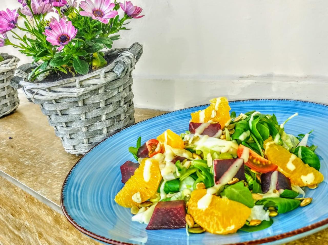 Kolorowa sałatka z pomarańczą, burakiem, pomidorkami cherry i pestkami dyni na niebieskim talerzu, obok wiklinowy koszyk z fioletowymi kwiatami.