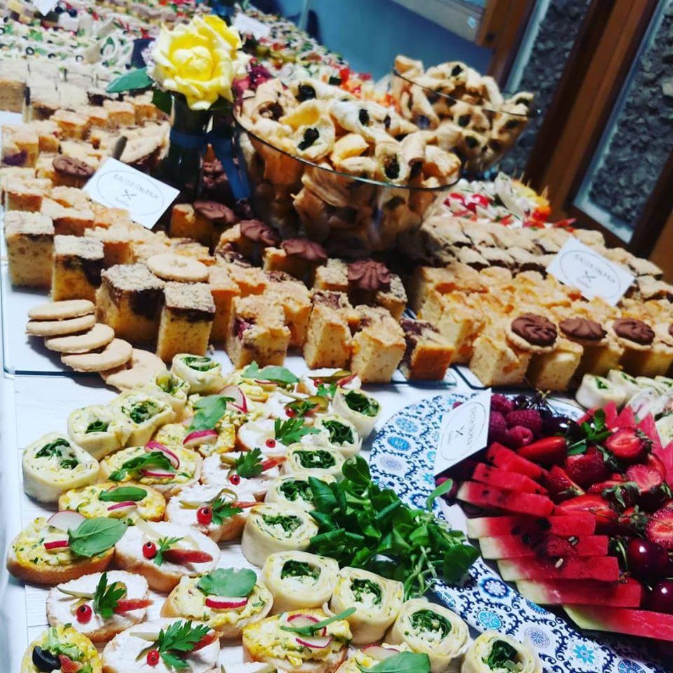 Bogato zastawiony stół z różnorodnymi przekąskami: ciasta w kostkach, ciasteczka, roladki z nadzieniem, kanapeczki z rzodkiewką i ziołami, arbuz, truskawki i wiśnie, dekoracja z żółtej róży w wazonie.