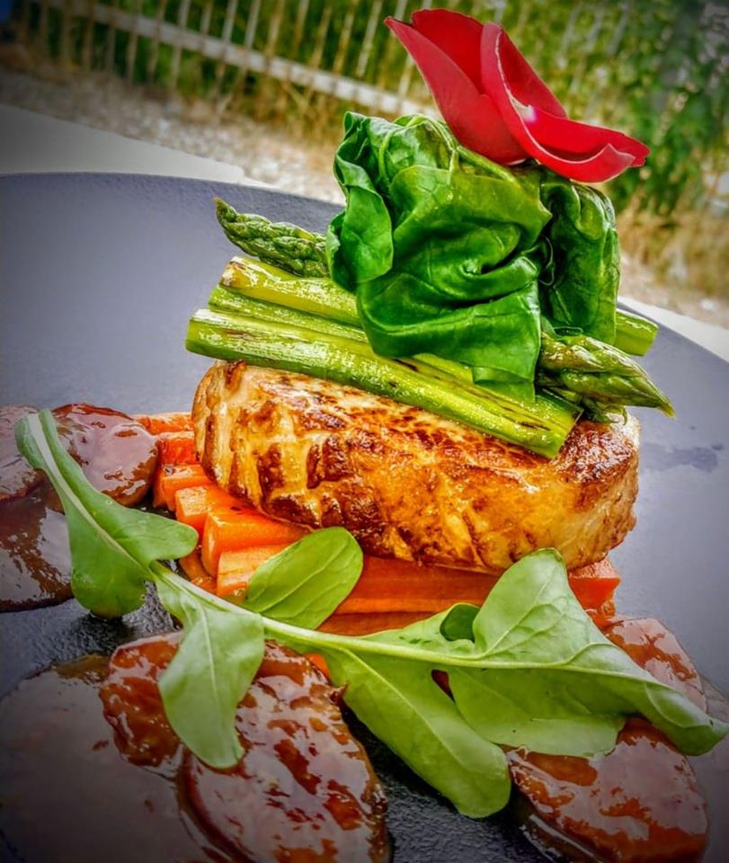 Elegancko podane danie: grillowany stek z warzywami, szparagami, sosem i dekoracją z płatków kwiatów na czarnym talerzu.