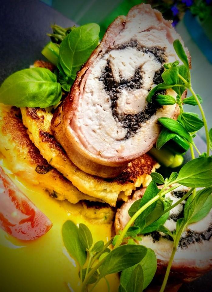 Rolada z jasnego mięsa z ciemnym farszem, otulona boczkiem, ułożona na plackach z sosem, udekorowana świeżymi ziołami i bazylią, na ciemnym talerzu.