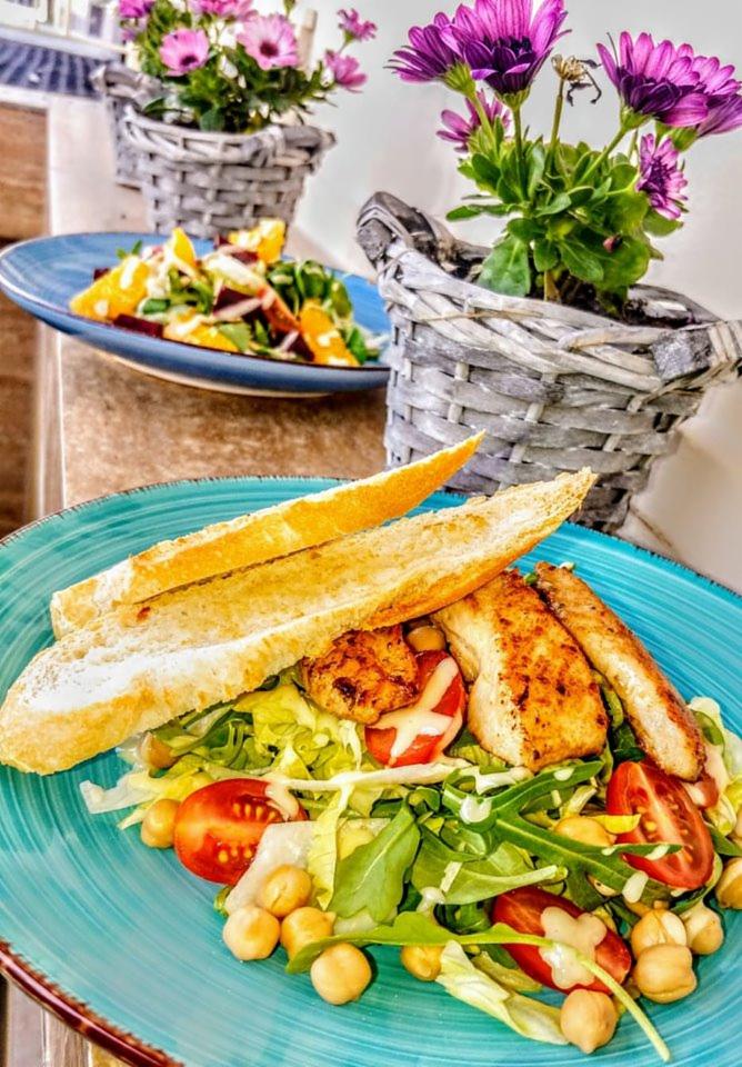 Turkusowy talerz z sałatką z grillowanym kurczakiem, grzankami, pomidorkami koktajlowymi, ciecierzycą i rukolą, w tle sałatka owocowa i kwiaty w wiklinowych koszykach.