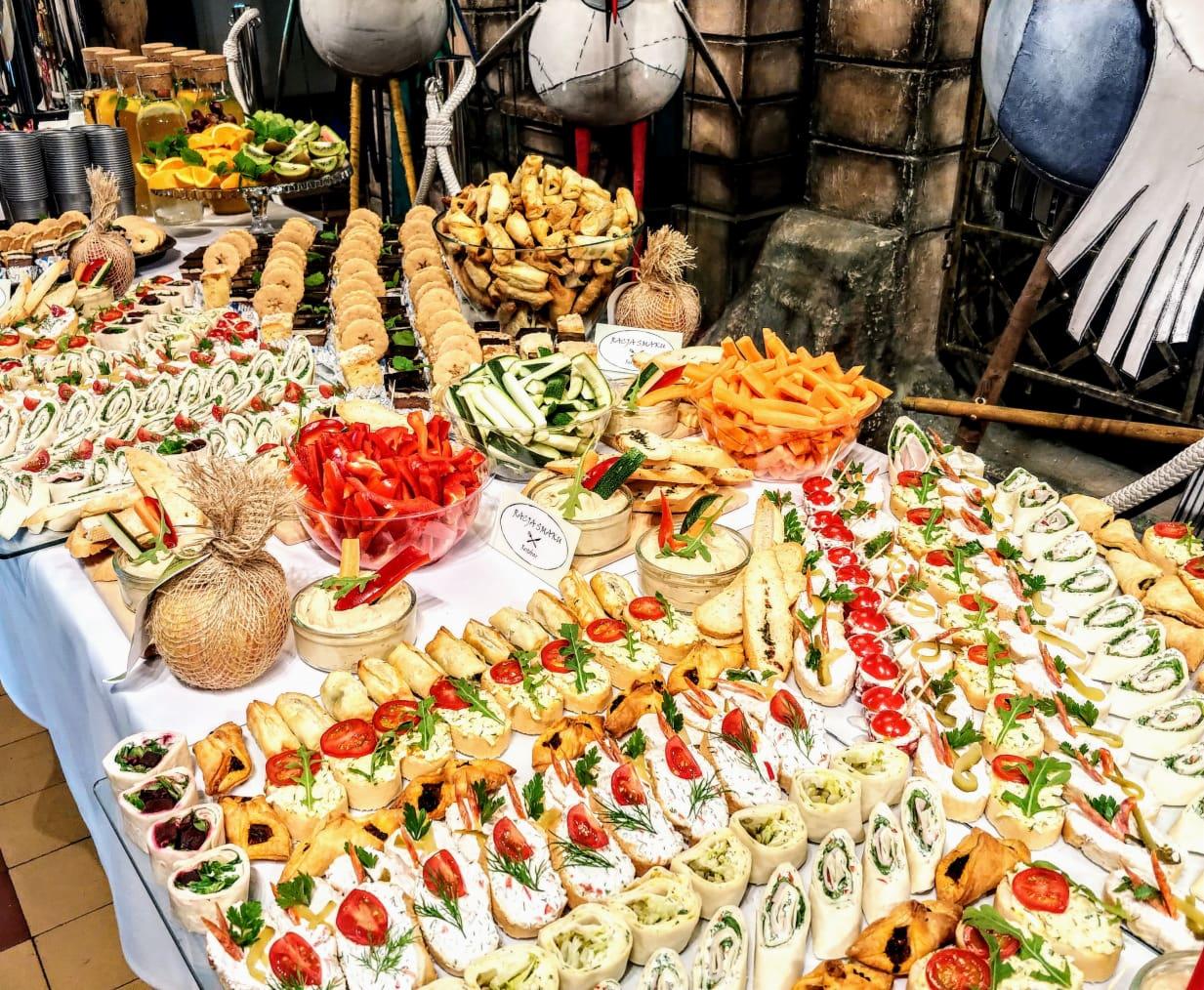 Bogato zastawiony stół z przekąskami: kanapeczki z pomidorami i ziołami, roladki z szynką i serem, warzywa, dipy, ciasteczka, soki; w tle dekoracje w stylu fantasy.