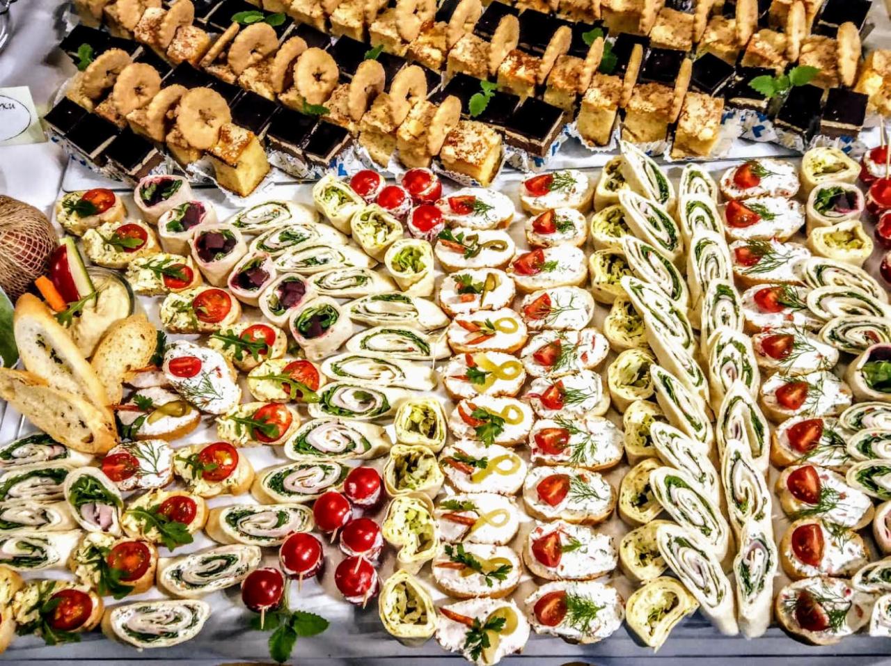 Bogaty wybór przekąsek na białym stole: ciasta, kanapki z pomidorami i ziołami, roladki z nadzieniem, bagietki i warzywa, gotowe do spożycia.