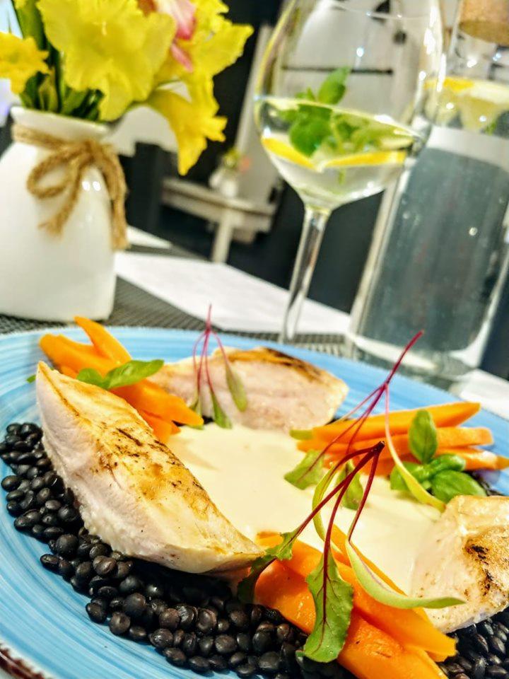 Elegancko podane danie z grillowanym kurczakiem, czarną soczewicą, marchewką i sosem na turkusowym talerzu, w tle wazon z żółtymi kwiatami i kieliszek wody z cytryną i miętą.