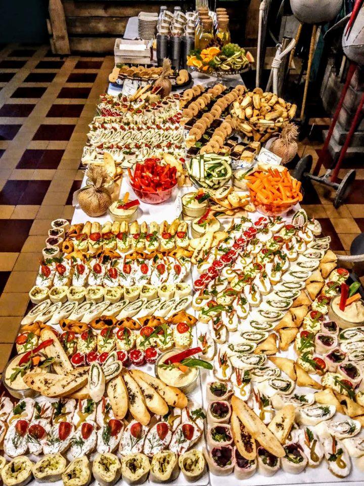 Bogato zastawiony stół z przekąskami w Krakowie, widok z góry. Różnorodność kanapek, tartinek i koreczków, z wyraźnie widocznymi pomidorami, ziołami i kremowymi pastami. W tle butelki z napojami...