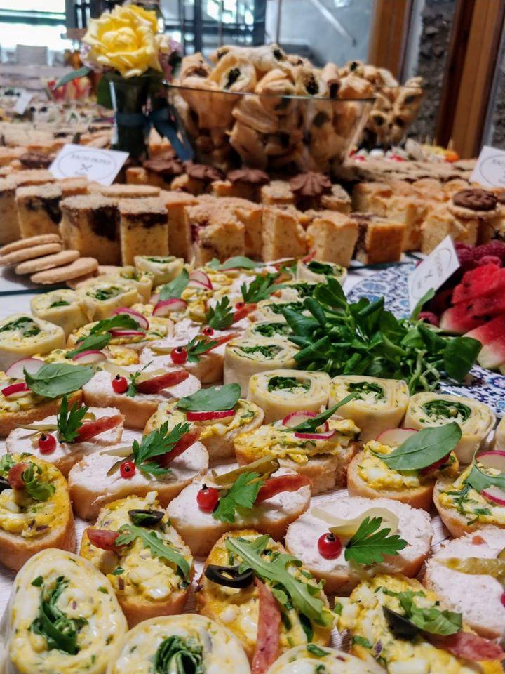 Bogato zastawiony stół z różnorodnymi przekąskami: mini kanapeczki z pastami, roladki szpinakowe, ciasta i ciasteczka, dekoracje kwiatowe, widoczne plasterki arbuza.