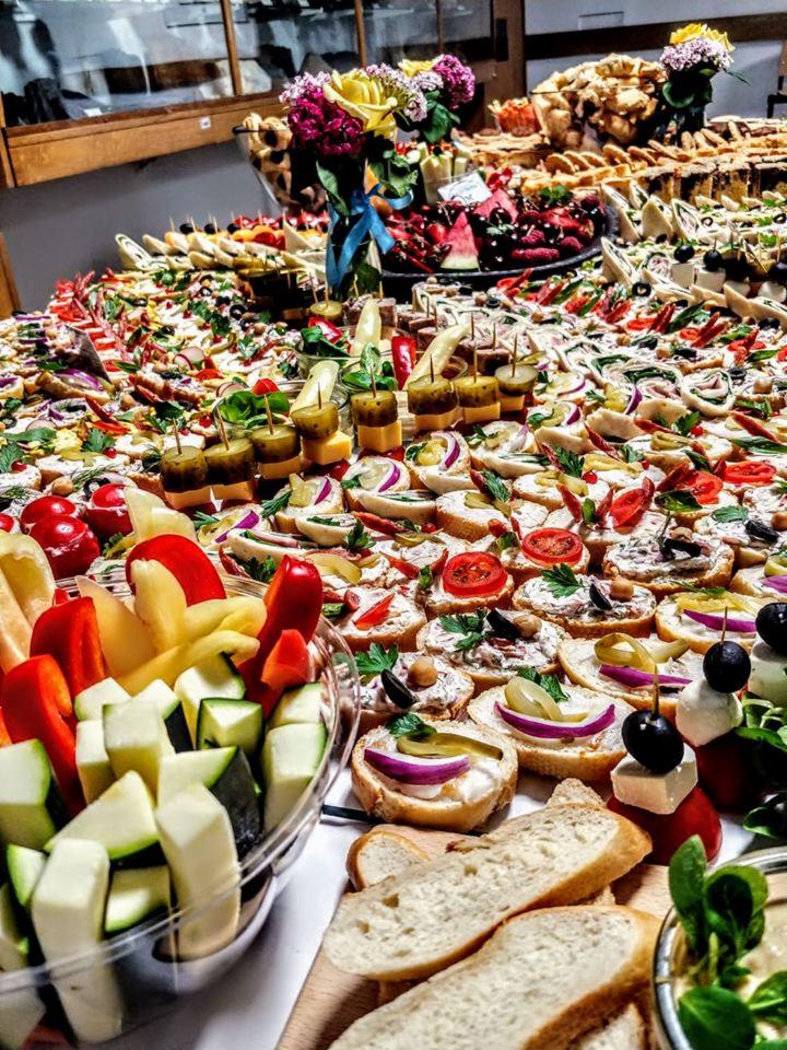 Stół obficie zastawiony różnorodnymi przekąskami: kanapeczki z warzywami, koreczki, pokrojone warzywa w misce, pieczywo, a w tle kompozycja kwiatowa.