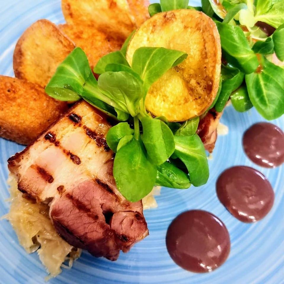 Delikatnie podpieczona karkówka z grilla na kapuście kiszonej, otoczona smażonymi ziemniakami, świeżymi liśćmi sałaty i trzema kroplami ciemnego sosu na niebieskim talerzu.