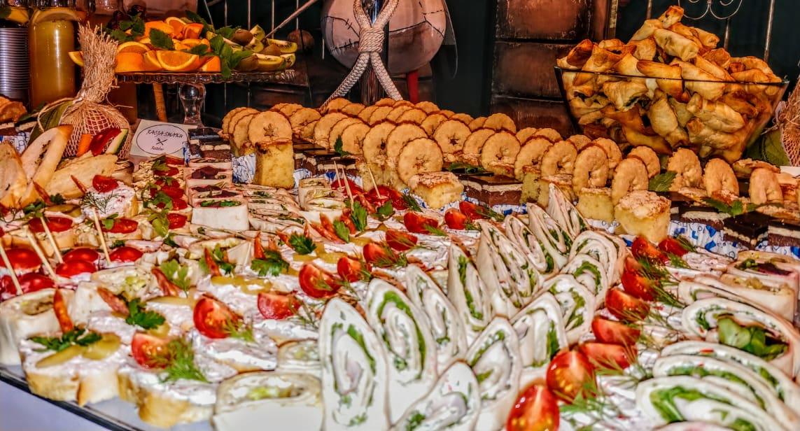 Bogaty stół z przekąskami: kanapki, roladki z nadzieniem, ciasta, owoce, soki i napoje, dekoracyjnie ułożone na przyjęciu.