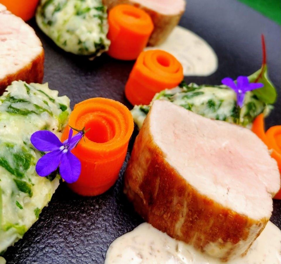 Elegancko podane danie z plastrami mięsa, puree ze szpinakiem, roladkami z marchewki i fioletowymi kwiatami na ciemnym talerzu z sosem.