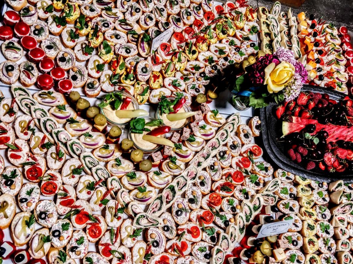 Bogato zastawiony stół z różnorodnymi przekąskami: mini kanapeczki z pomidorami, oliwkami i ziołami, roladki z szynką i serem, hummus z warzywami w szklanych naczyniach oraz taca z owocami...