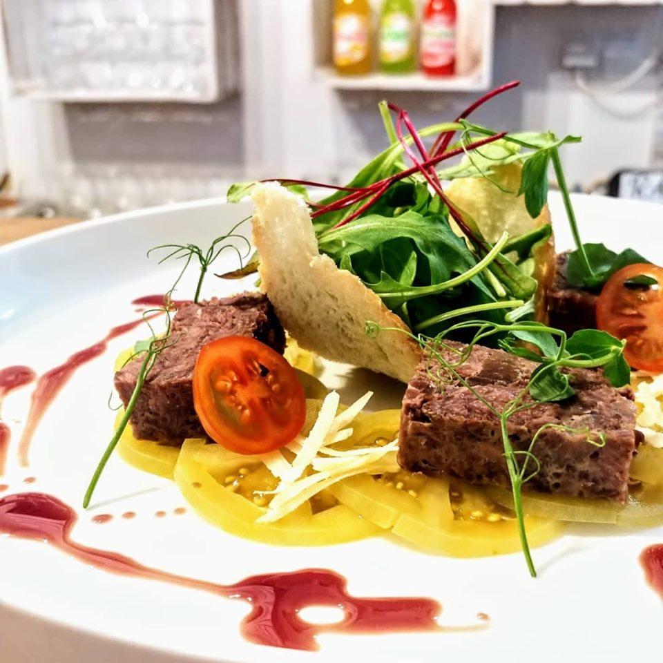 Wykwintna przystawka na białym talerzu: pasztet z żółtymi pomidorami, rukolą i sosem malinowym, dekorowana mikro-zielenią i kawałkiem chleba, z butelkami napojów w tle.