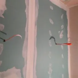 Ściana z płyt kartonowo-gipsowych w trakcie montażu, widoczne kable elektryczne wystające ze ściany, szpachlowane łączenia płyt i otwory po wkrętach.