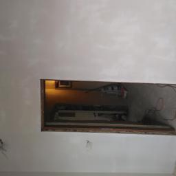 Dominik Muracki - Wnętrze w trakcie remontu: ściany po nałożeniu pierwszej warstwy gładzi, widoczne niedoskonałości i ślady po szpachlowaniu, przygotowany otwór w ścianie z widocznymi przewodami i narzędziami...